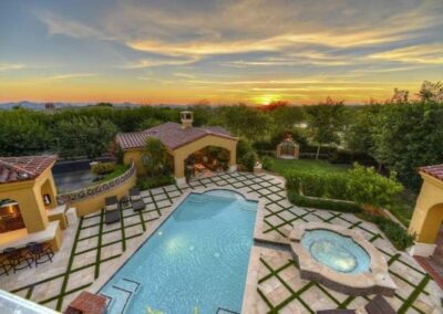 Painted desert Sky over landscape featuring custom pavers surrounding a pool, outdoor grill, multiple covered seating areas, and well-maintained trees and shrubs.