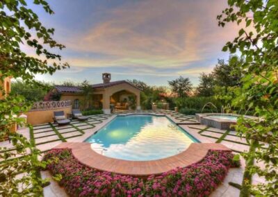 Red flowering bushes surrounding a pool with custom pavers, grass between the stones, tree maintenance, and elegant water features.