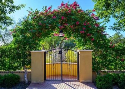 Flowering vines arching over an entry gate, with manicured hedges lining a brick wall that conceals a long, elegant driveway.